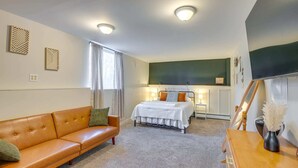 Una habitación de hotel con un sofá de cuero marrón, una cama con ropa blanca, un escritorio con un jarrón y un ventanal con cortinas traslúcidas.