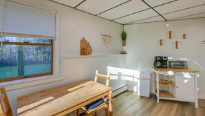 Cocina compacta con una mesa y sillas de madera, un refrigerador pequeño, cafetera y tostadora. Una ventana con persianas deja entrar luz natural, y una planta añade un toque de verdor.