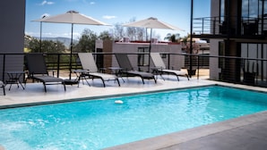 Una piscina con sillones de descanso y sombrillas, rodeada por un edificio moderno con grandes ventanales.