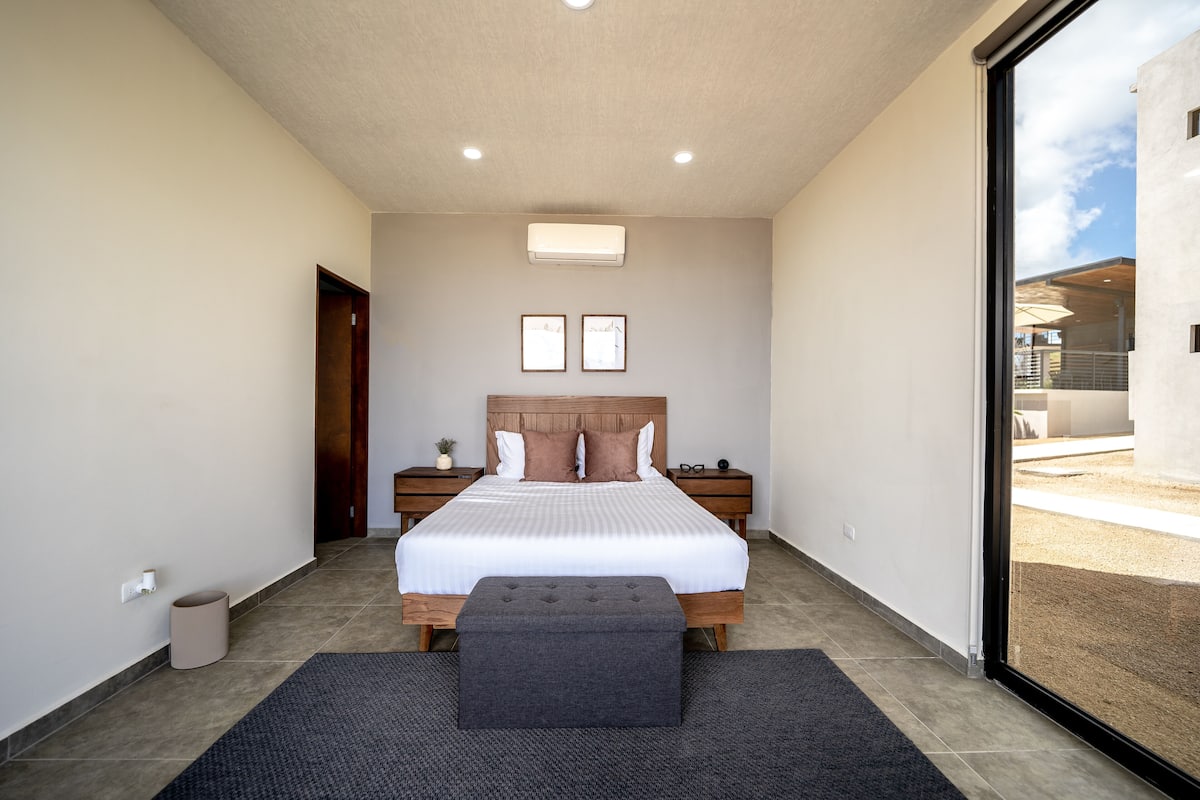 A bedroom with a large bed, a bedside table, a small stool, and a window with a view of the outside.