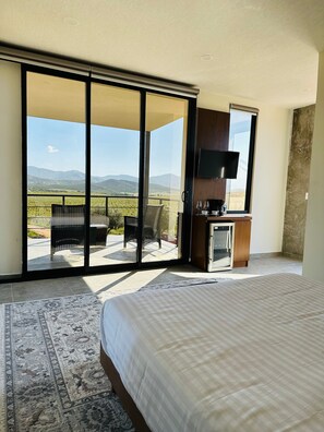 Een slaapkamer met een groot bed, een televisie, een kitchenette en een schuifpui naar een balkon met uitzicht op het platteland.