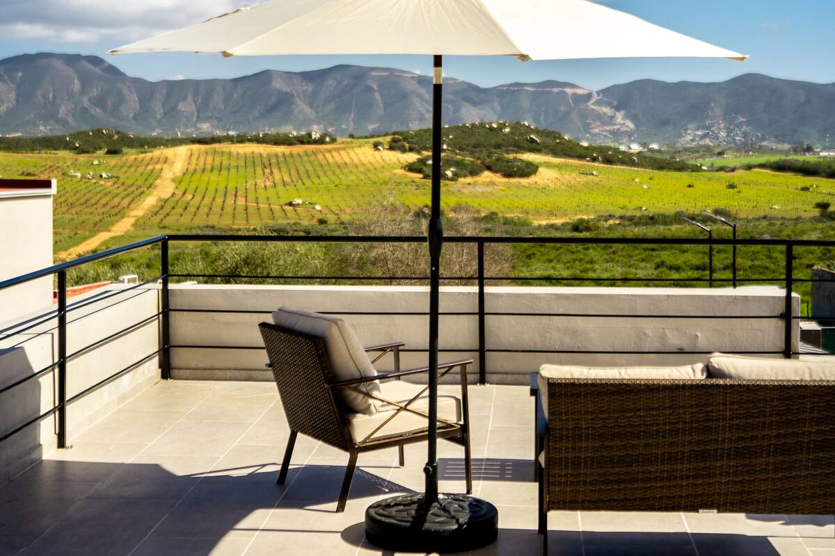 A terrace with a wicker sofa, a table, and an umbrella, overlooking a green landscape with mountains in the distance.