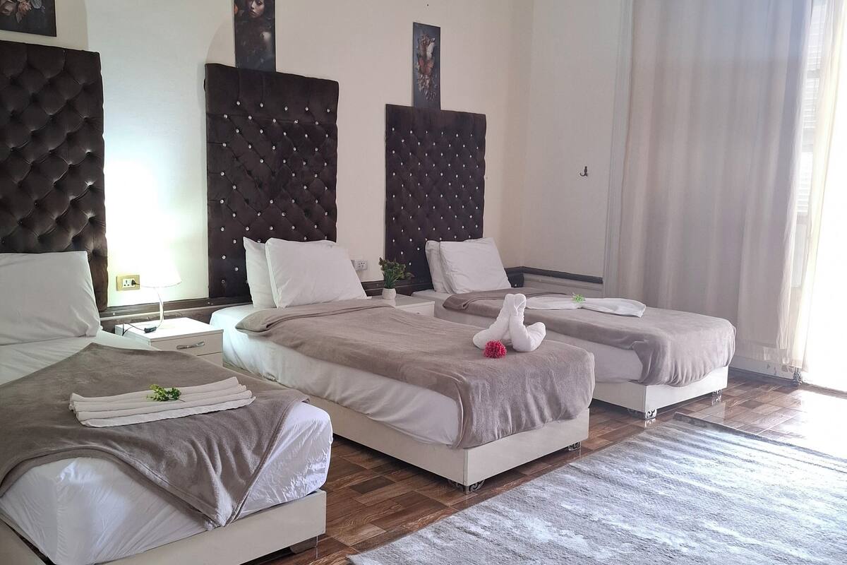 A hotel room with two beds, a large window with curtains, a wooden floor, and artwork on the walls.