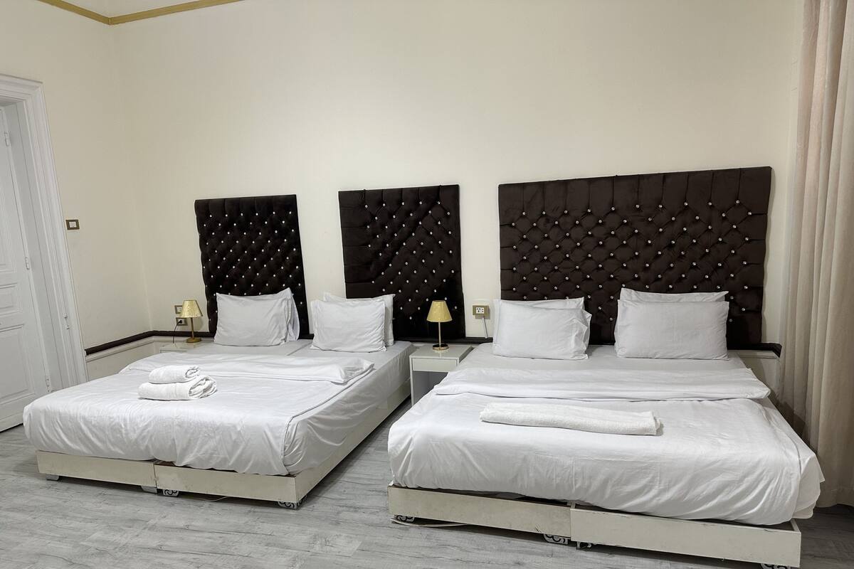 A hotel room with two beds, white bedding, a dark tufted headboard, a nightstand with a lamp, and a wooden floor.