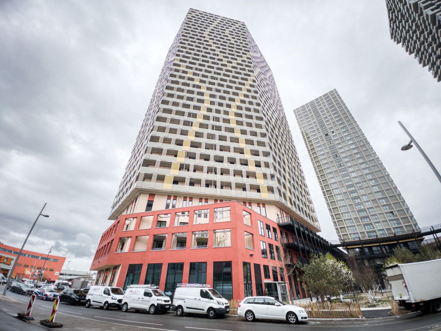 Un immeuble de grande hauteur à la façade quadrillée, situé à côté d’un bâtiment rouge doté de nombreuses fenêtres, avec des véhicules stationnés devant.