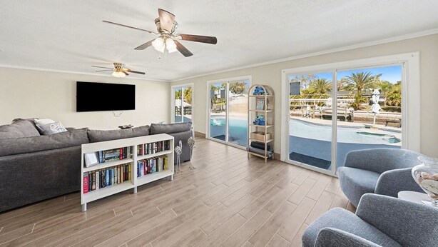 A spacious living room with a grey sofa, a white bookshelf, and a ceiling fan. Large windows offer a view of a swimming pool and palm trees. - Bayside Retreat with Backyard Oasis (Tampa)