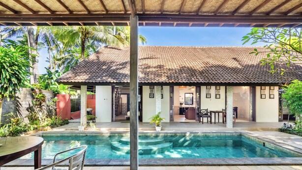 A pool area with a covered patio, a dining area with tables and chairs, and a view of a building with a tiled roof. - Surya Villa (Karangasem)