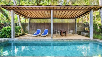 A pool area with a covered seating area, two blue lounge chairs, and a table with chairs.