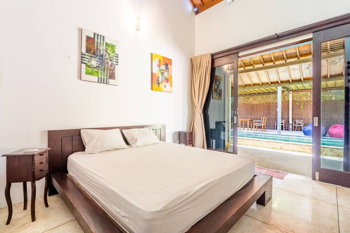A bedroom with a large bed, a nightstand, and a sliding glass door leading to an outdoor area with a pool.