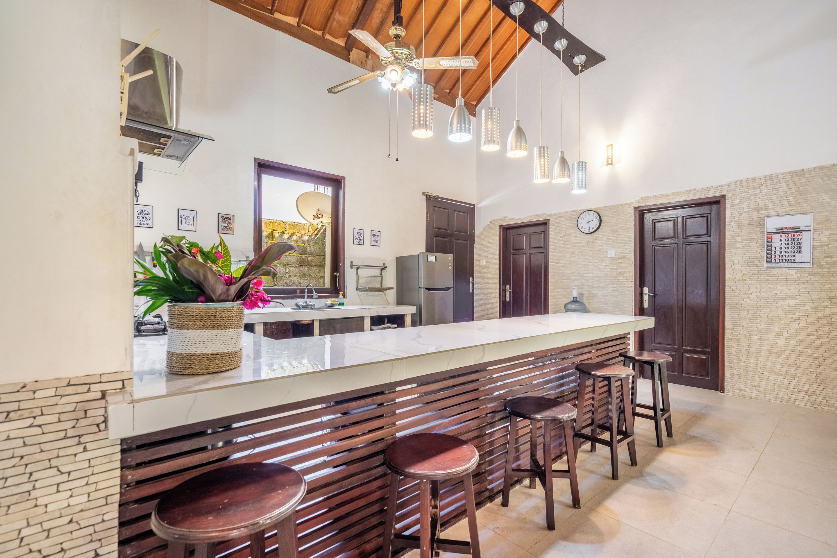 Una cocina con barra, taburetes de madera, ventilador de techo, luces colgantes y pared de ladrillo.