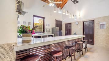 Una cocina con barra, taburetes de madera, ventilador de techo, luces colgantes y pared de ladrillo.