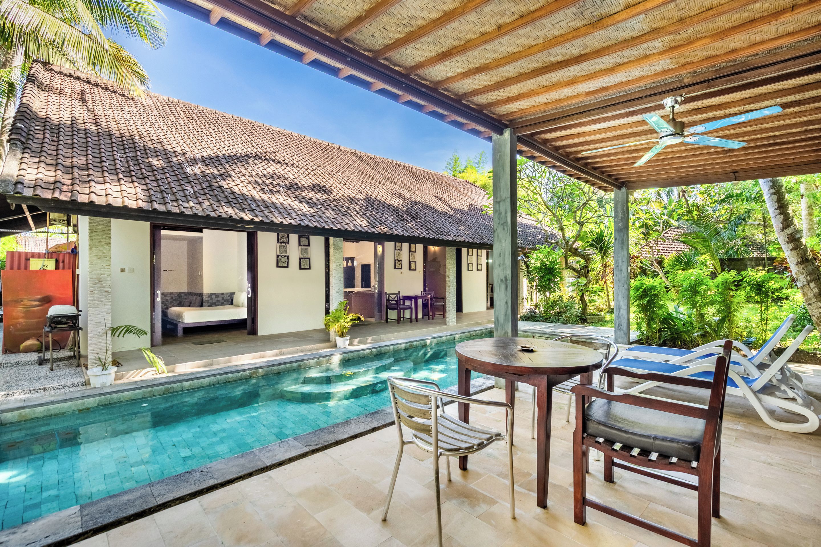 Zona junto a la piscina con área de descanso cubierta, una mesa y sillones reclinables.