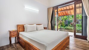 A bedroom with a wooden bed, a nightstand, and a view of a garden through a sliding glass door. - Surya Villa (Karangasem)