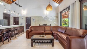 A modern living area with a leather sectional sofa, a wooden coffee table, and a kitchen with a bar area. - Surya Villa (Karangasem)
