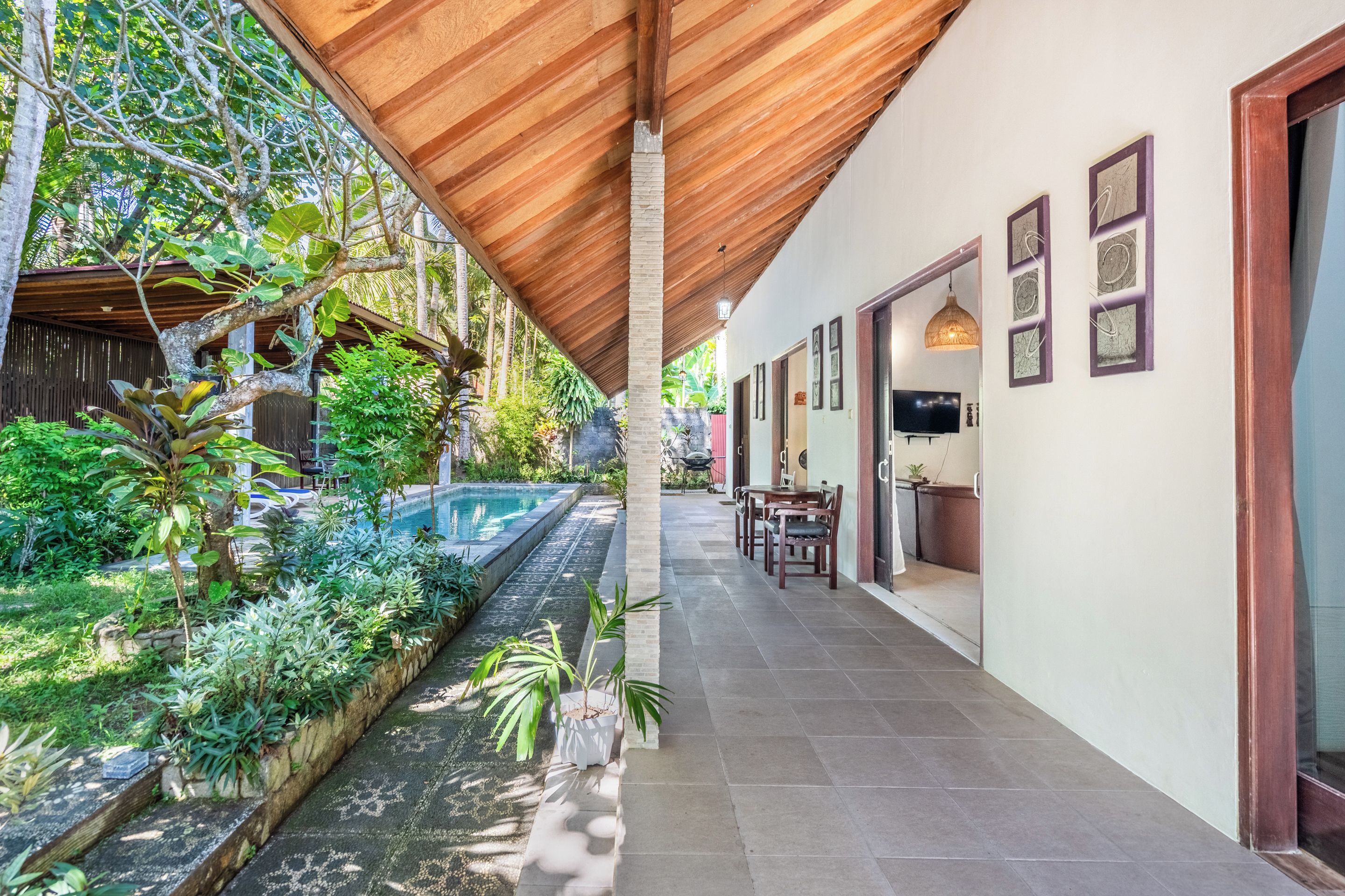 A covered outdoor area with a pool, surrounded by lush greenery and a dining area with a TV.