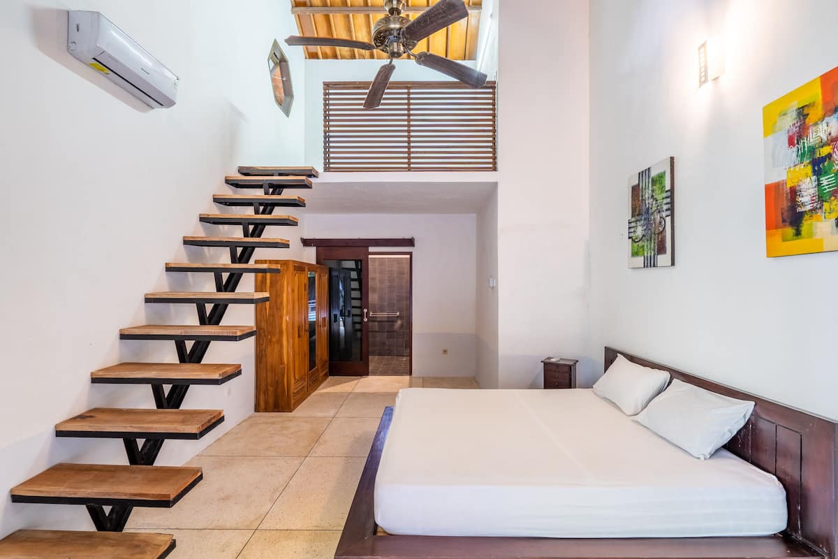 A bedroom with a bed, wooden staircase, ceiling fan, and artwork on the wall.
