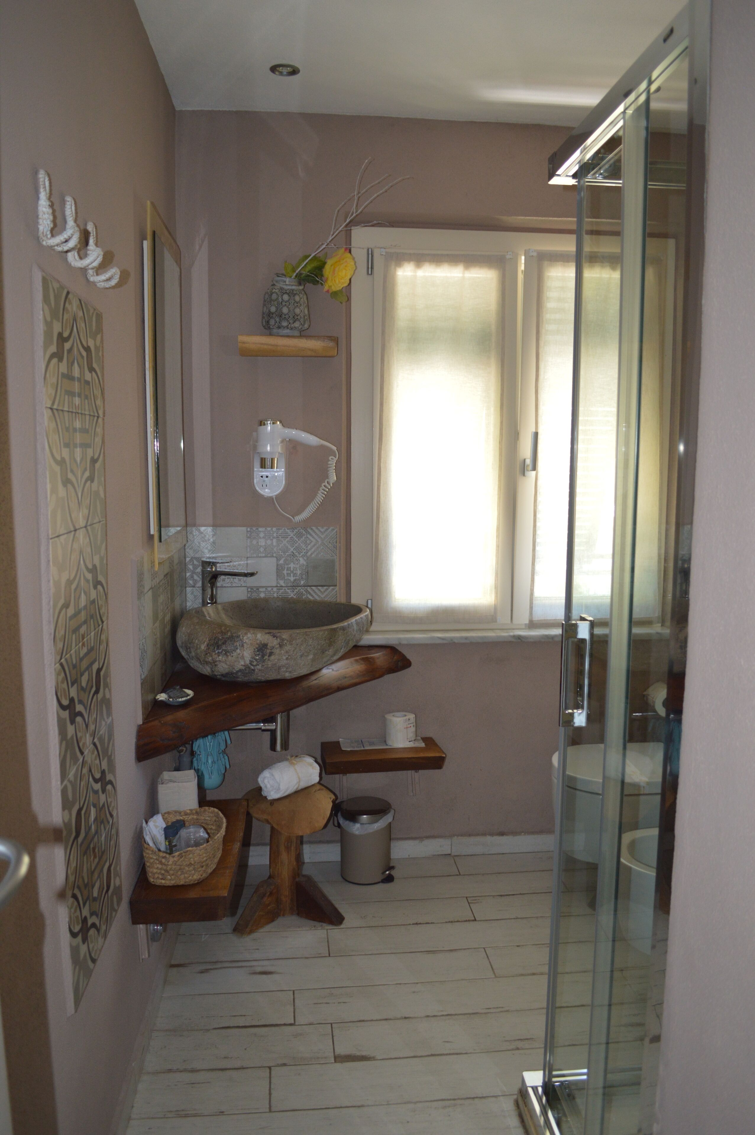 A bathroom with a stone sink, a wooden shelf, a glass shower enclosure, and a window with blinds.