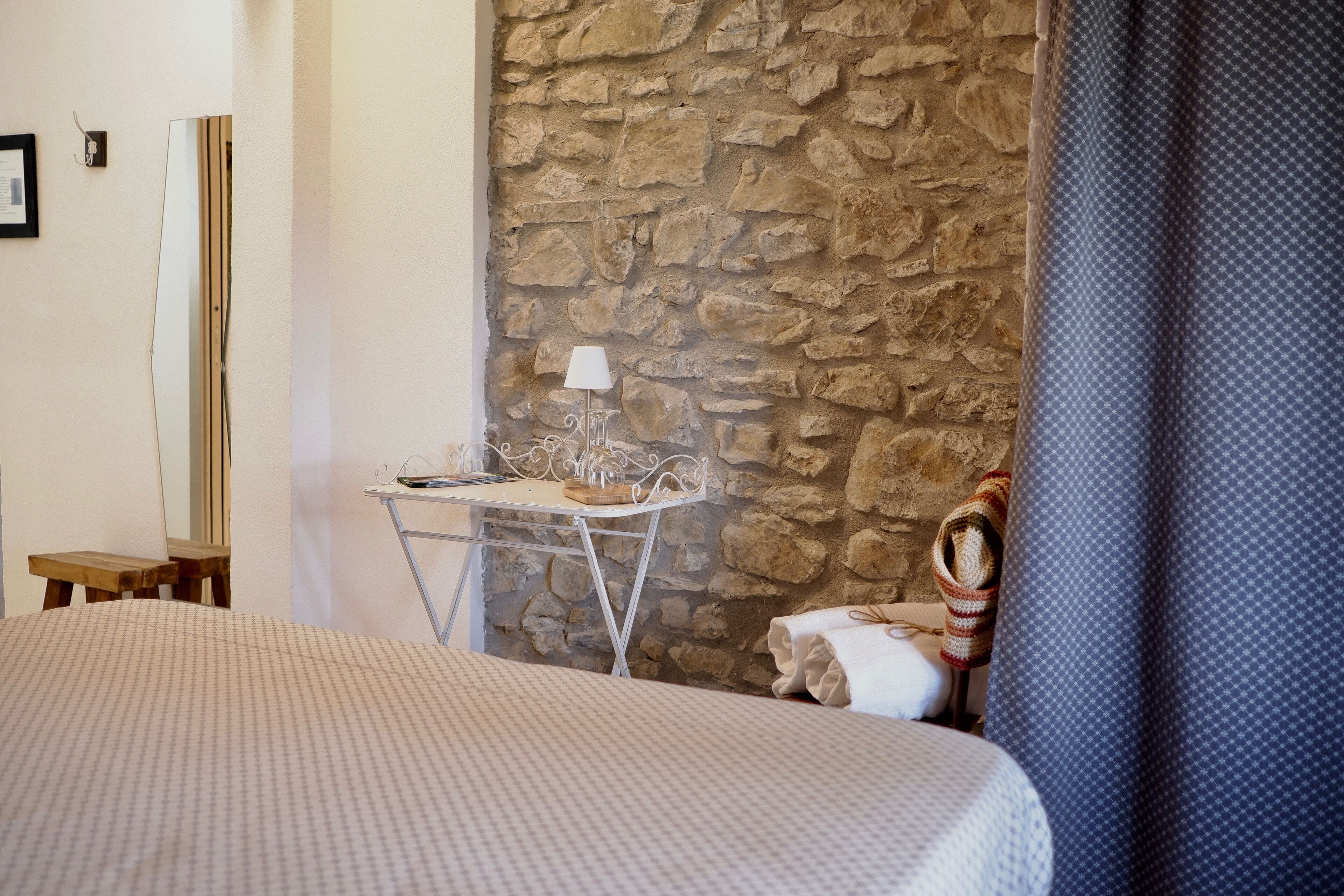 A bedroom with a bed, a small table with a lamp, a stone wall, and a window with a curtain.