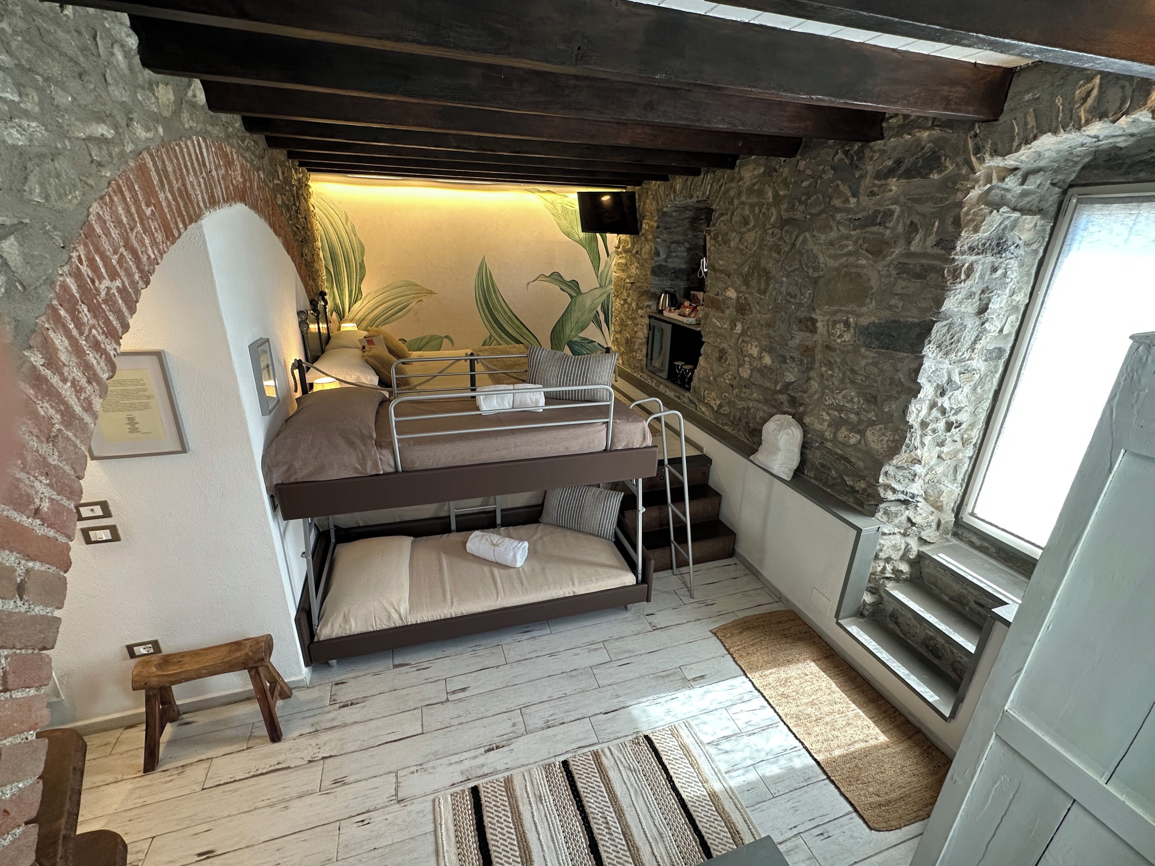 Una habitación con una cama individual, un pequeño banco de madera, un muro de piedra, una ventana y una planta.