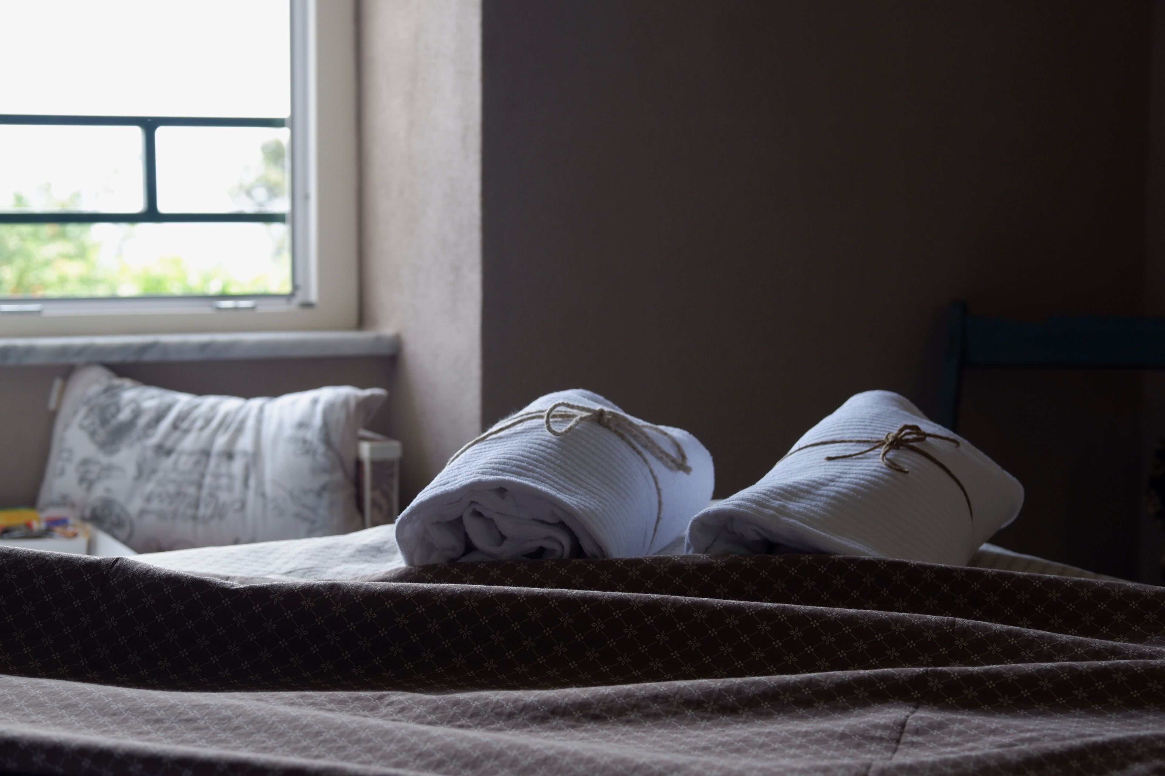 Un lit bien fait, avec des serviettes pliées et un oreiller, une fenêtre donnant sur un paysage verdoyant et une chaise au fond de la pièce.
