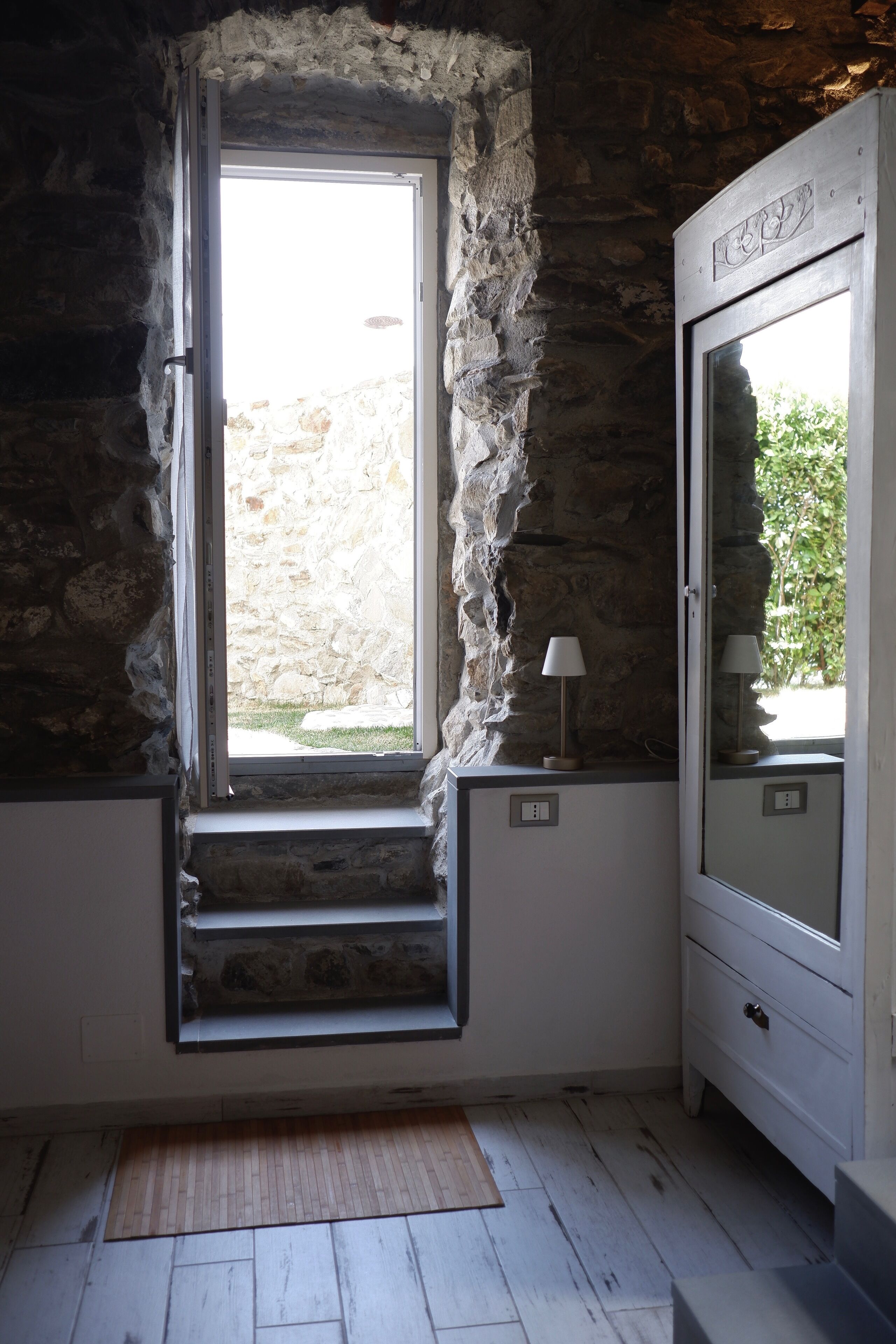 A room with a stone wall, a window with a view, a small table with a lamp, and a white cabinet.