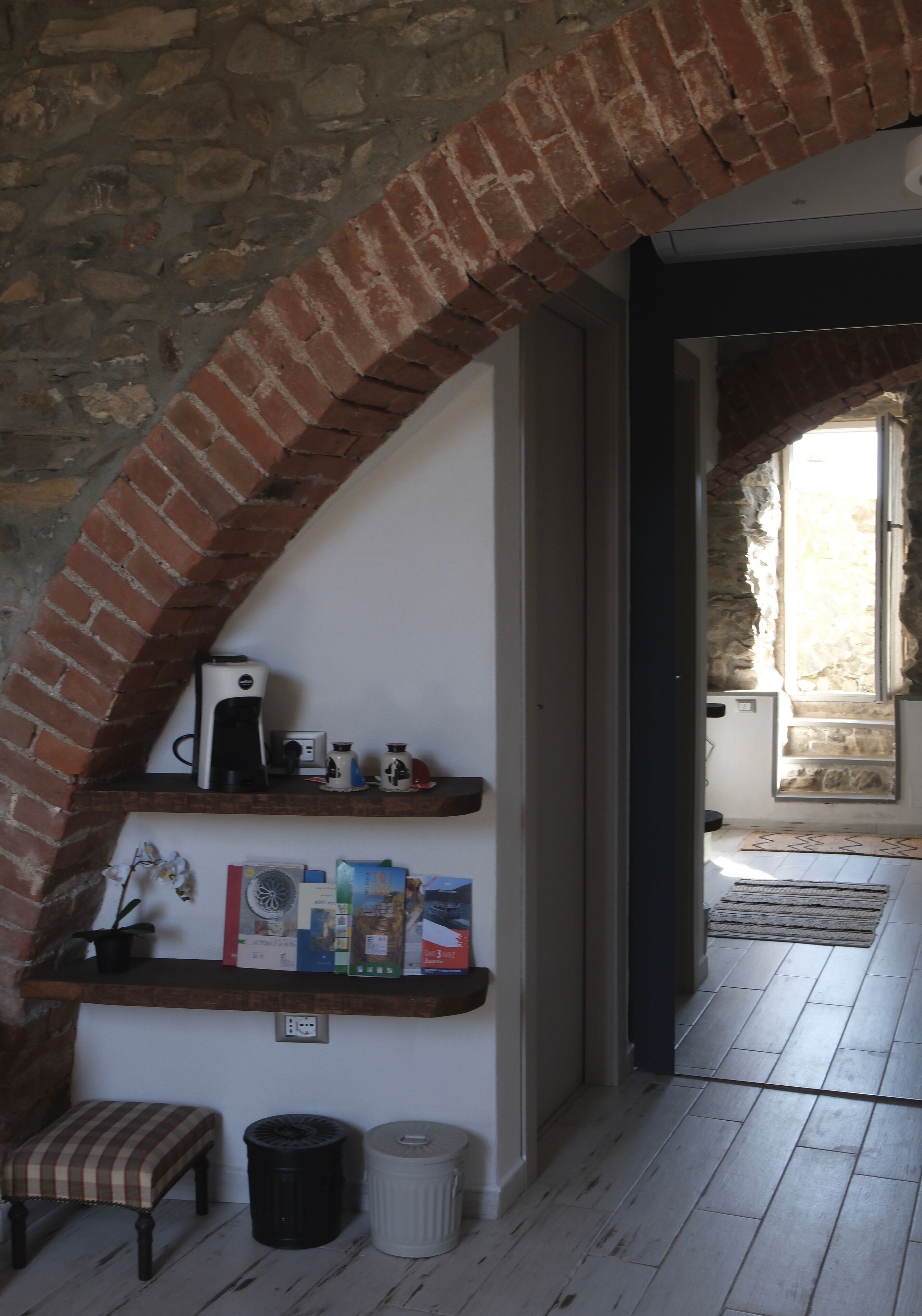 A cozy room with a brick archway, a shelf with books and a kettle, a bench, and a trash bin.