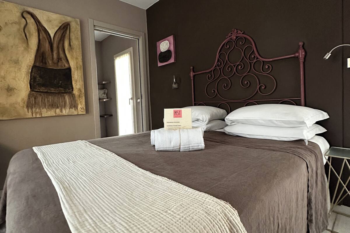 A neatly made bed with a headboard, a bedside table, a wall-mounted clock, and a painting on the wall.