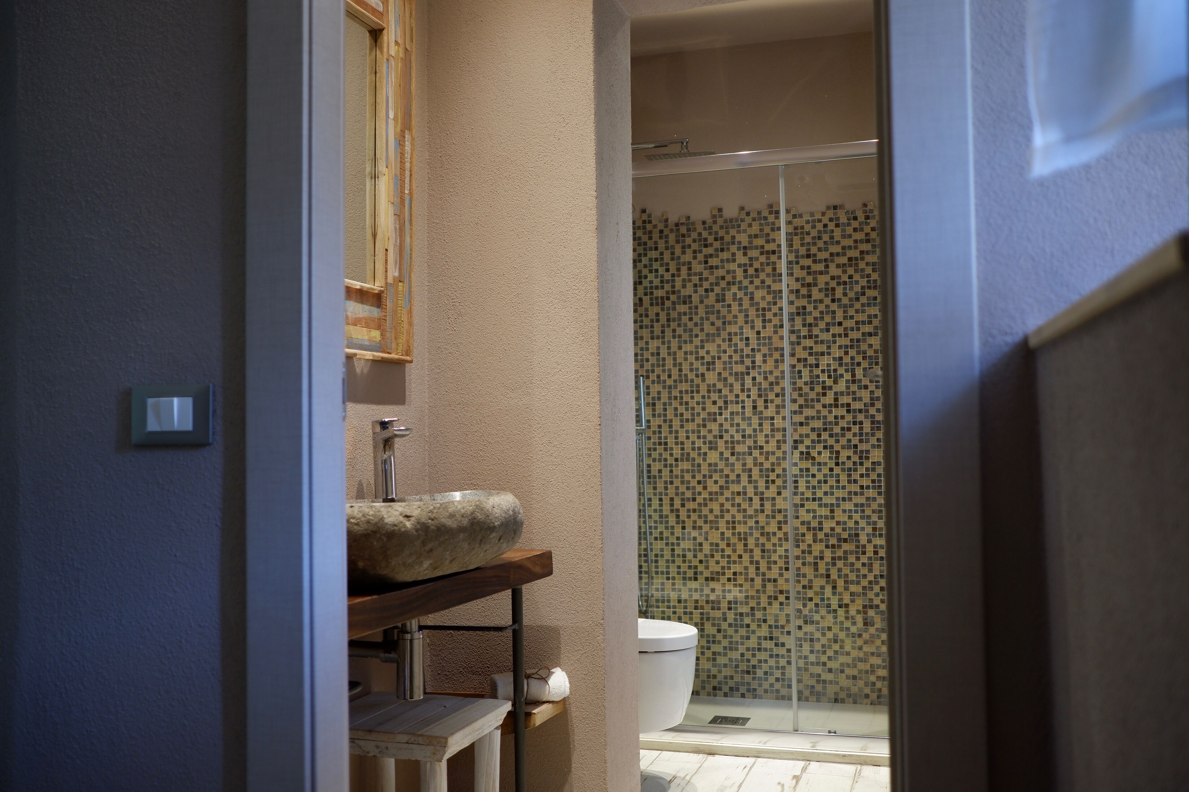 A bathroom with a stone sink, a wooden shelf, a toilet, and a shower with mosaic tiles.