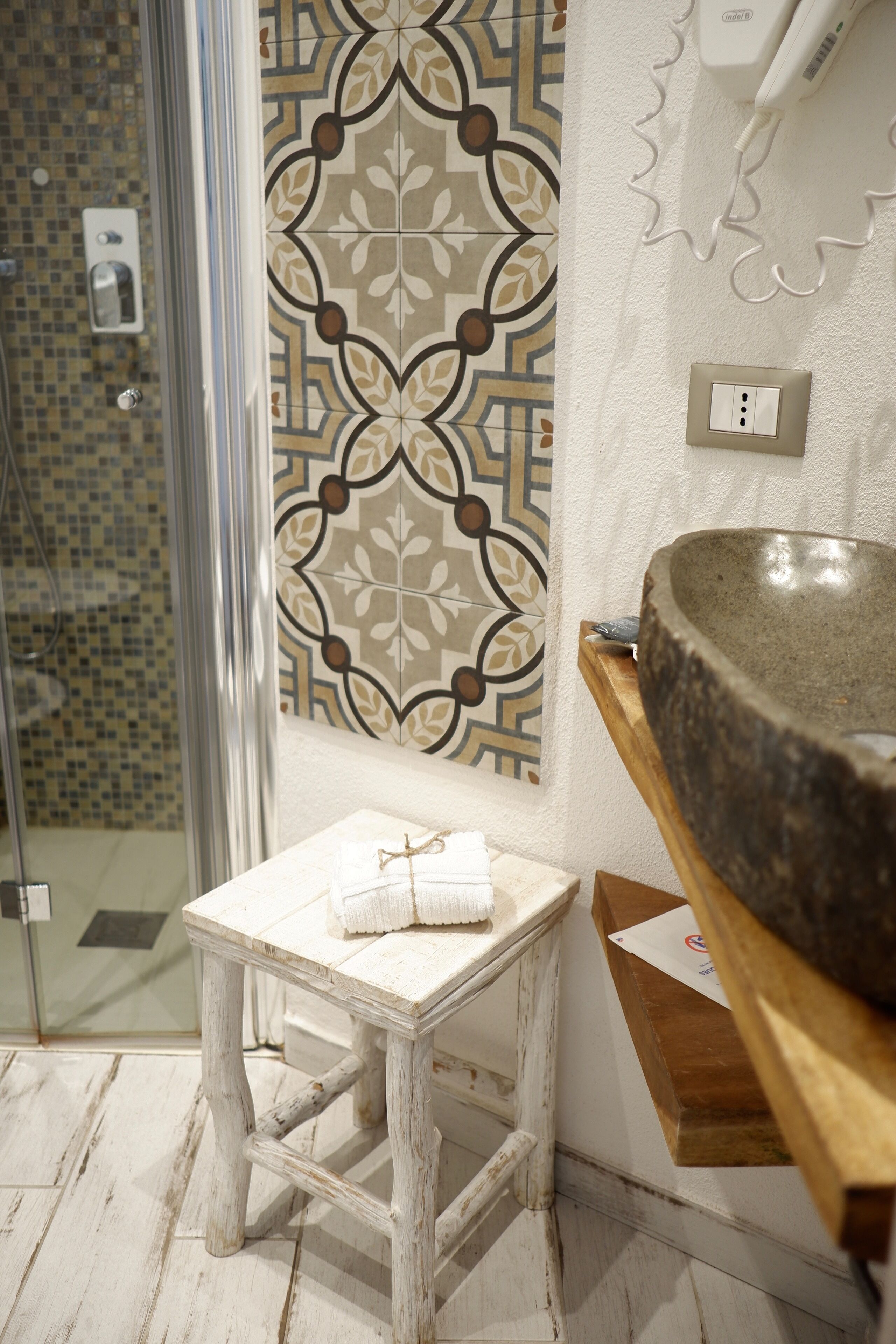 A bathroom with a glass shower door, a patterned tile wall, a wooden stool with a towel, and a stone sink.