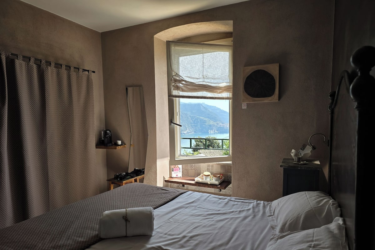 A bedroom with a bed, a window with a view of mountains, a shelf with items, and a small table with a lamp.