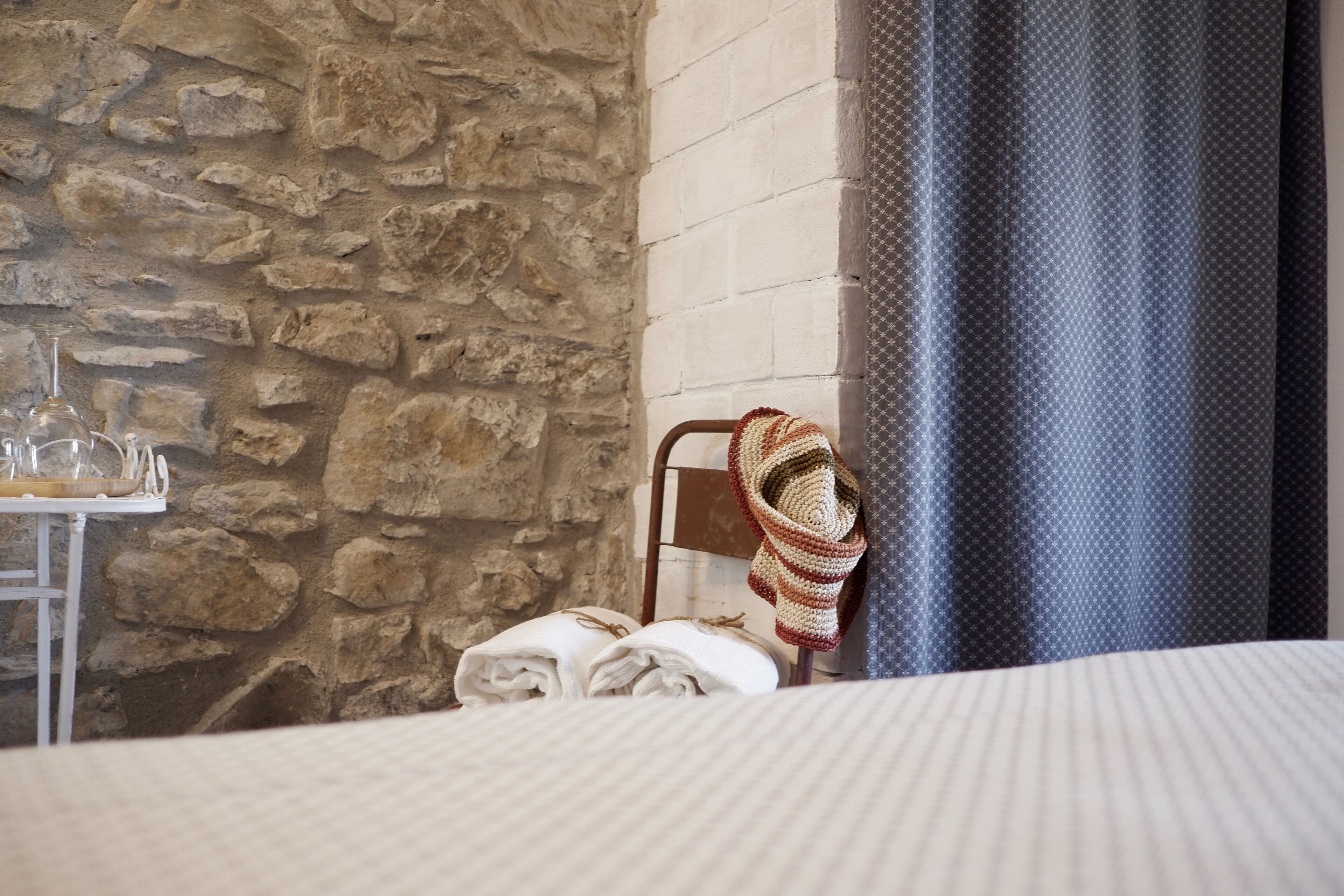 A hotel room with a stone wall, a bed with a striped bedspread, a chair with a knitted blanket, and a table with a glass set.