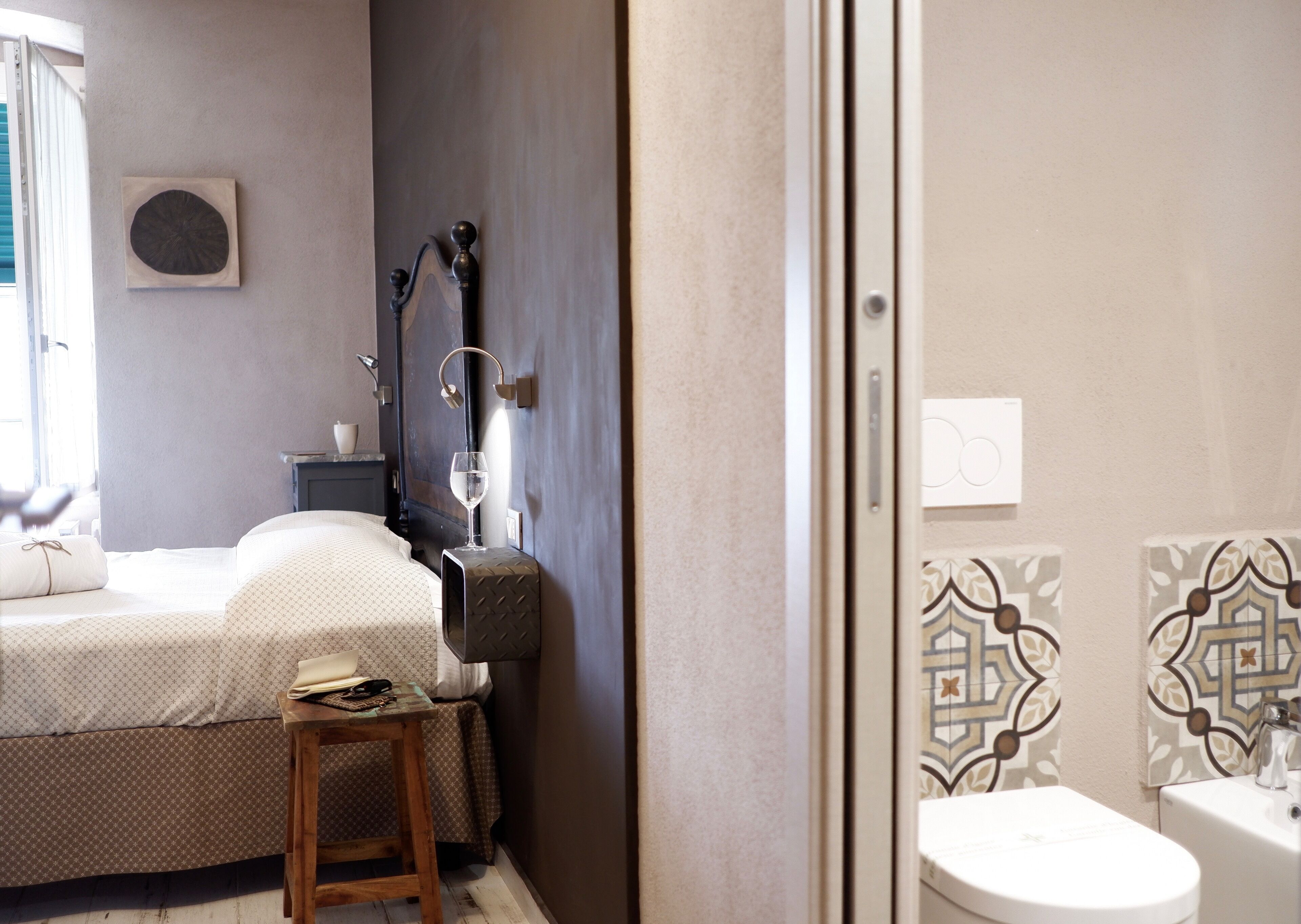Un dormitorio con cama, mesita de noche y lámpara de noche. Hay una ventana con persianas, un reloj de pared y una lámpara de pared.