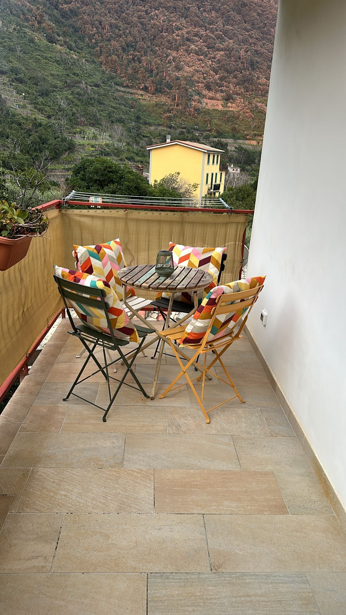 A balcony with a table and chairs, a potted plant, and a view of a yellow building in the background.