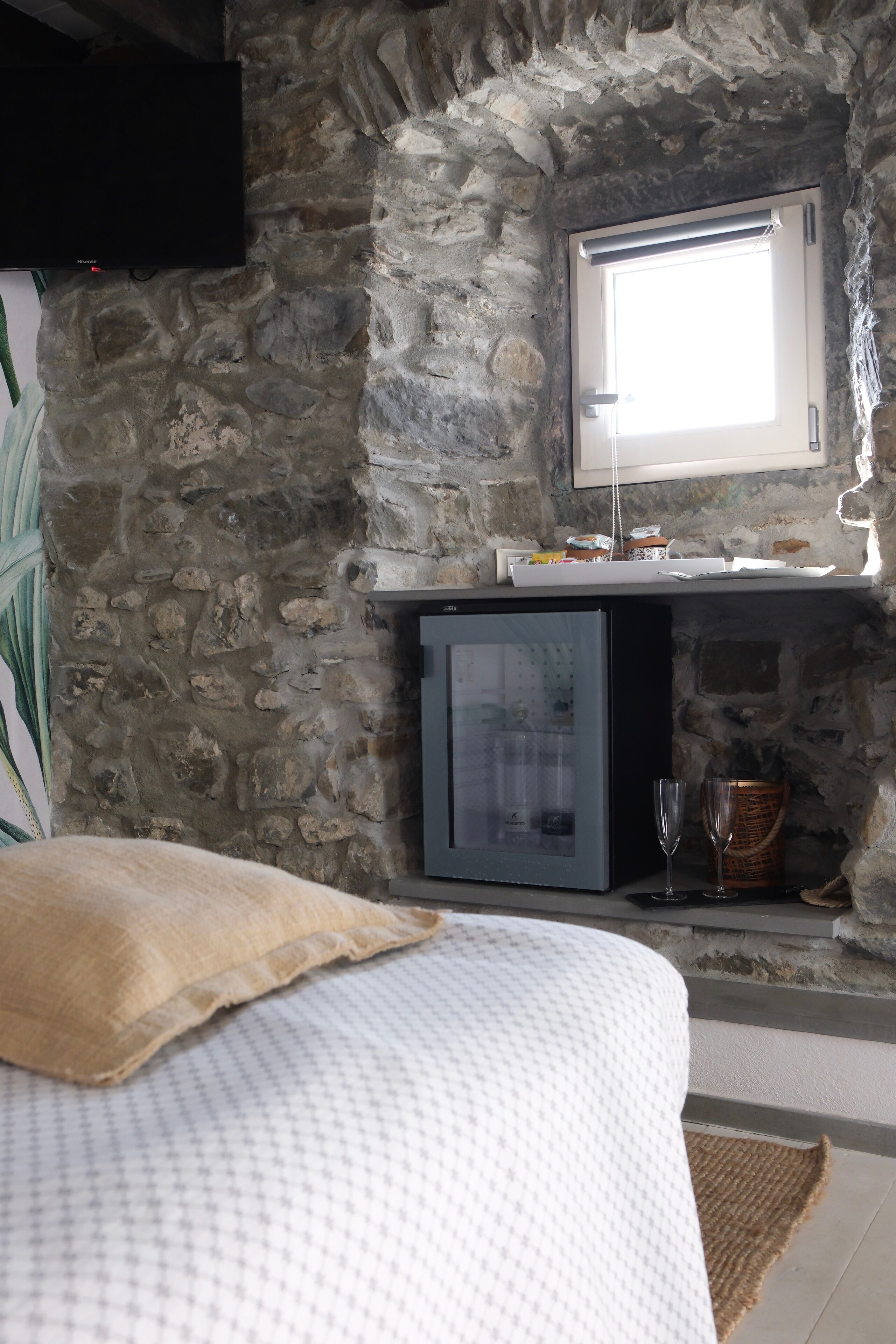 A stone-walled room with a bed, a television mounted on the wall, and a window.