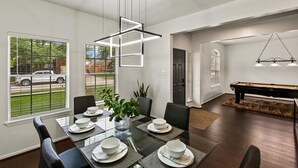 Une salle à manger moderne avec une table en verre dressée pour quatre personnes, entourée de chaises noires. Un luminaire suspendu se trouve au-dessus de la table, et une table de billard est visible au fond.