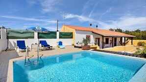 Ein Swimmingpool mit blauem Wasser, ein überdachter Bereich mit Liegestühlen und ein Gebäude mit rotem Dach.