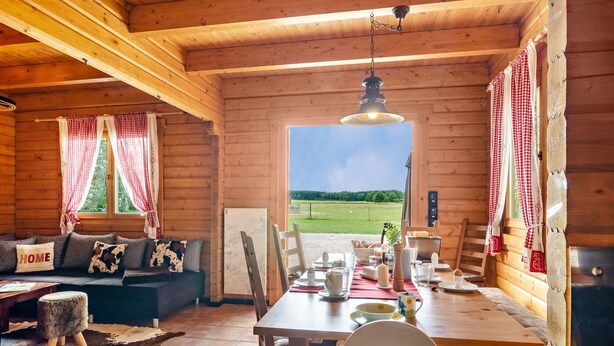Interior de una cabaña de madera con sofá, mesa de comedor y vistas al exterior.