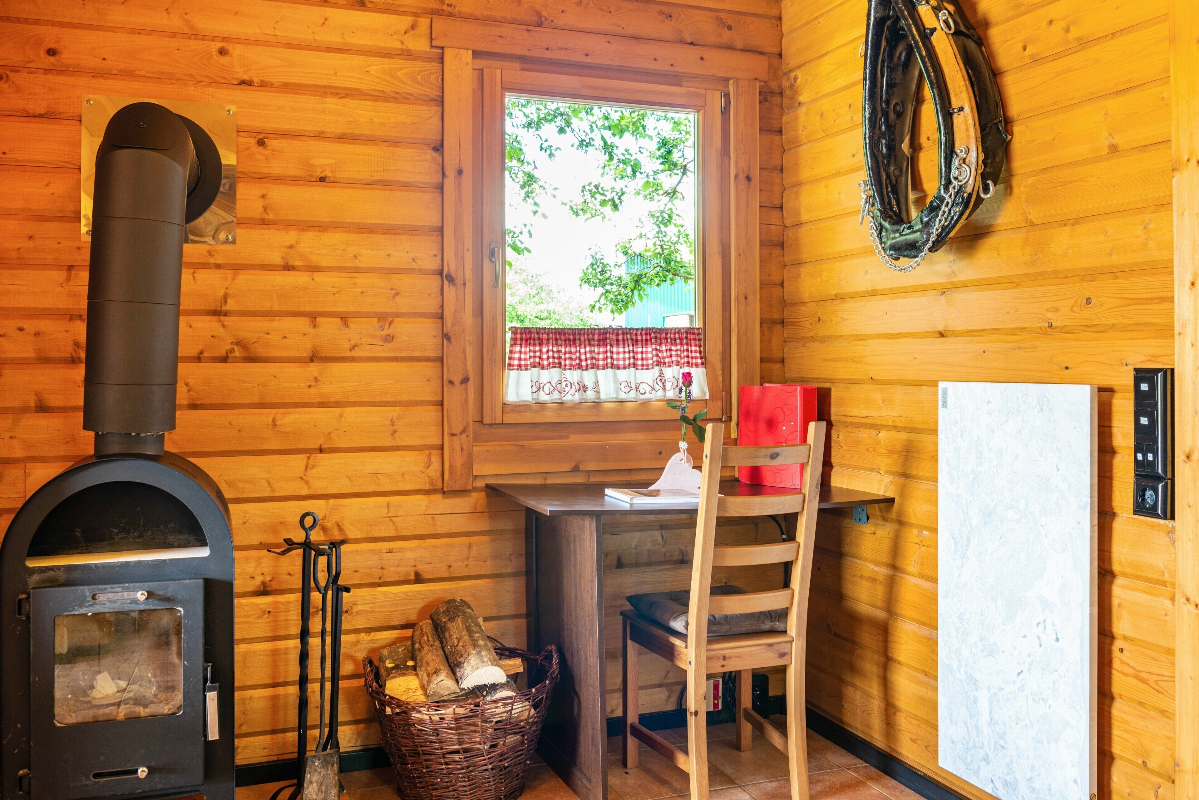 Interior de una cabaña de madera con un estufa de leña, una ventana con un cubre ventana rojo y blanco a cuadros, una silla de madera y una cesta de leña.