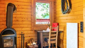 Interior de una cabaña de madera con un estufa de leña, una ventana con un cubre ventana rojo y blanco a cuadros, una silla de madera y una cesta de leña.