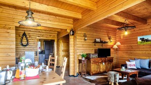 Interior de una cabaña de madera con mesa de comedor, sillas, sofá, televisor y zona de cocina.