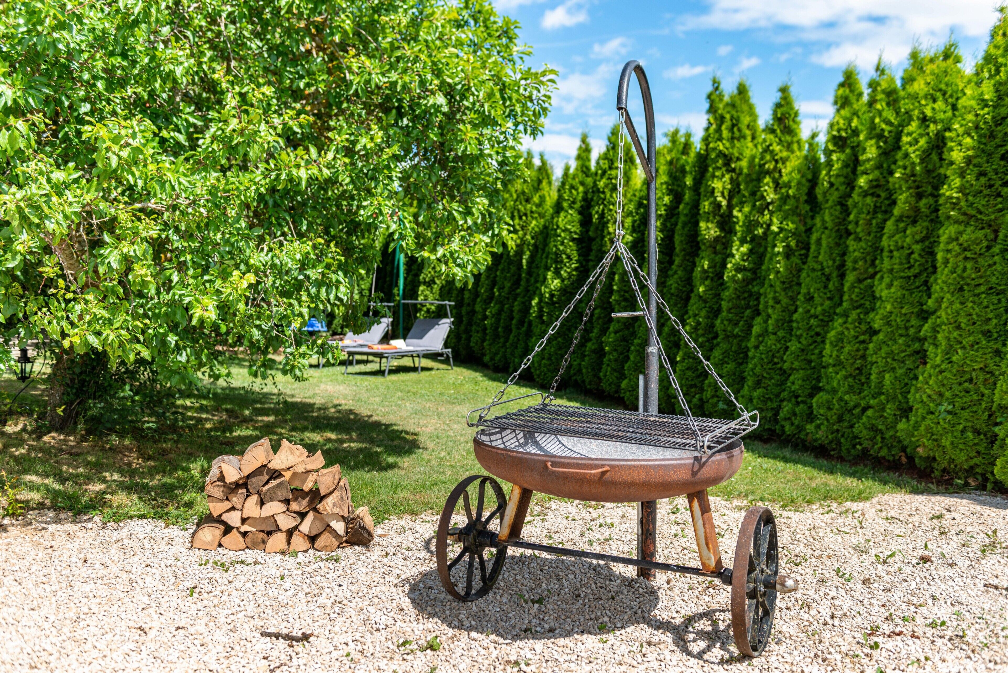 A metal grill with a chain and wheels, a pile of wood, and a chair in a garden.