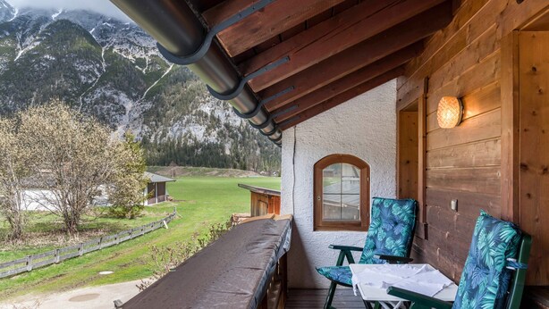 A wooden balcony with a table and chairs, a window, and a view of a mountainous landscape.