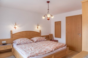 A bedroom with a wooden bed, bedside tables, a nightstand, a wall-mounted lamp, a wardrobe, and a door.