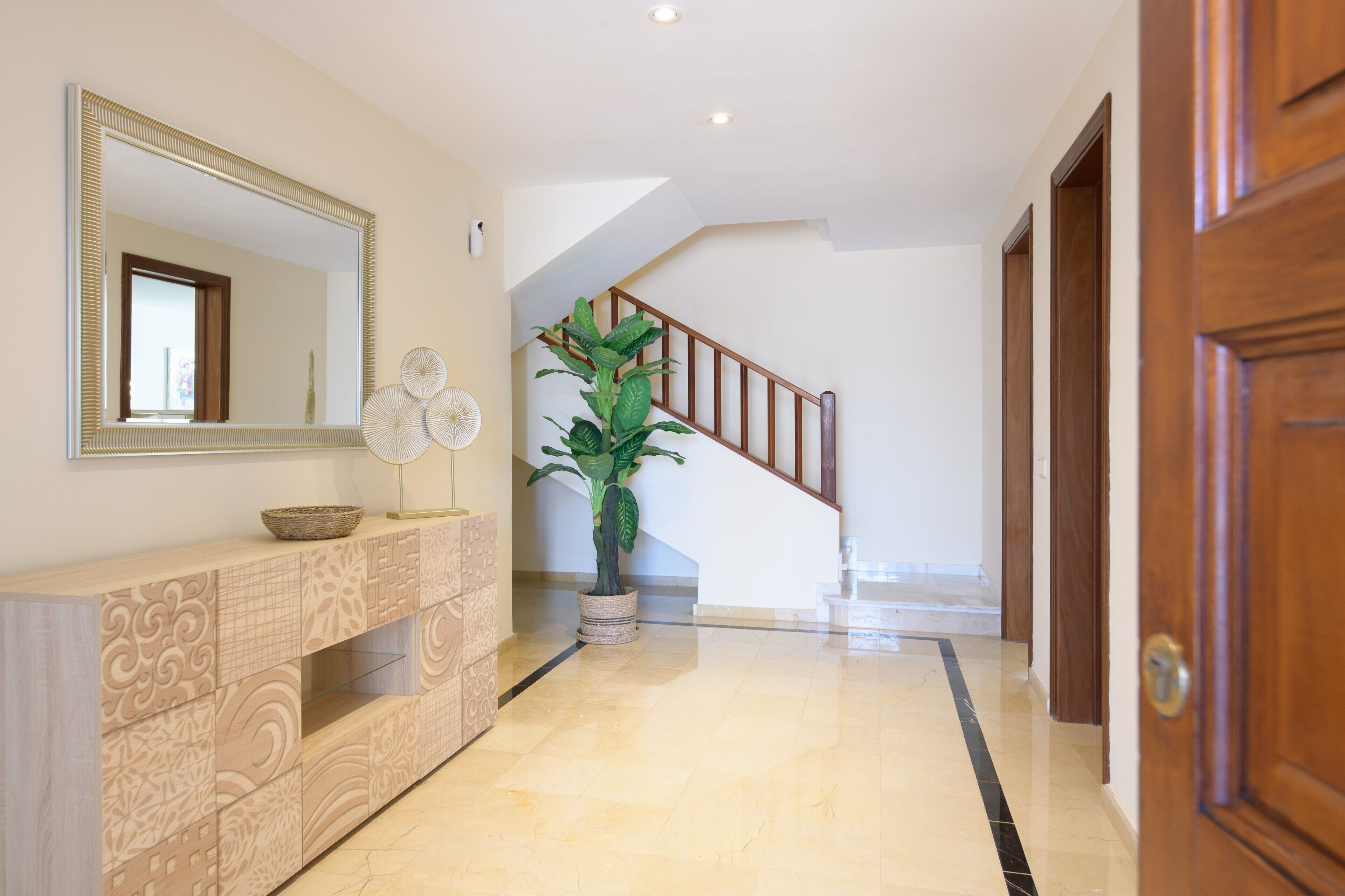 Un pasillo con una escalera de madera, un espejo grande, una consola decorativa y una planta en maceta.