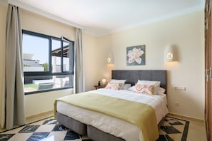 A hotel room with a bed, a window with curtains, a wall-mounted light, and a painting.