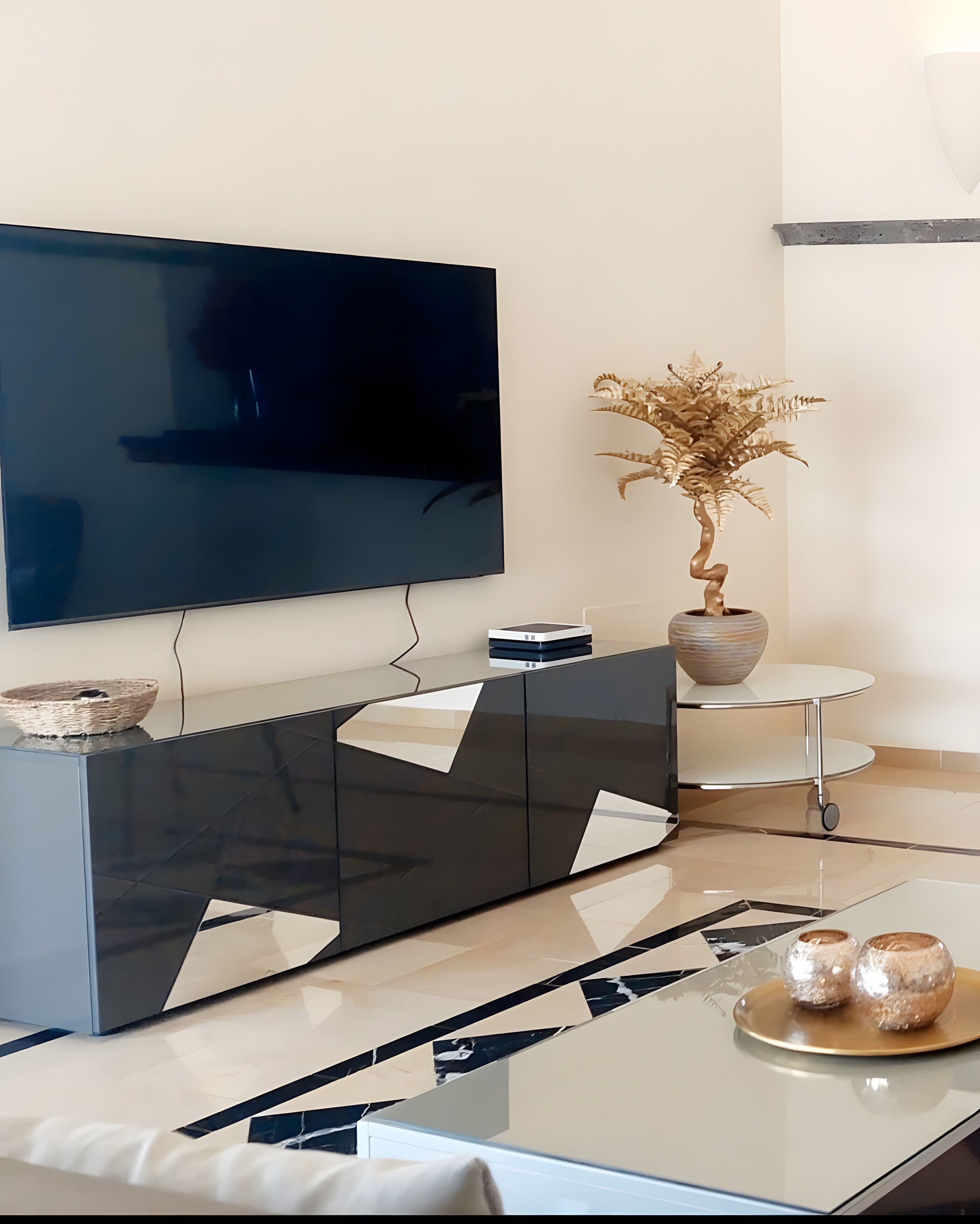 Ein modernes Wohnzimmer mit Flachbildfernseher an der Wand, einem eleganten schwarz-weißen Schrank, einer Topfpflanze und einem Glastisch mit Dekoration.