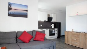 Ein modernes Wohnzimmer mit grauem Sofa, roten Kissen, einer Küchenzeile mit weißen Schränken und einem Landschaftsbild.