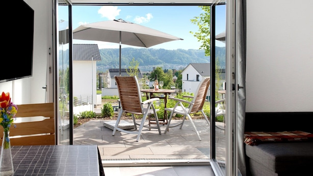 Eine geöffnete Schiebetür, die auf eine Terrassenanlage mit Tisch, Stühlen und Sonnenschirm führt.