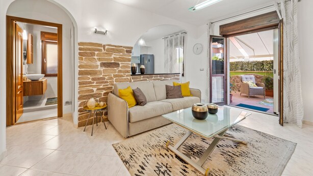 A modern living room with a stone wall, a sofa, a glass coffee table, and a view through a sliding glass door. - Holiday Home "Ca' Alessandra" with Private Terrace and Air Conditioning (Carloforte)