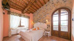A bedroom with a stone wall, a wooden ceiling, a bed with pillows, a nightstand, and a window with curtains. - Villa Podere Cafaggio with Private Pool, Terrace, and Air Conditioning (Monteverdi Marittimo)